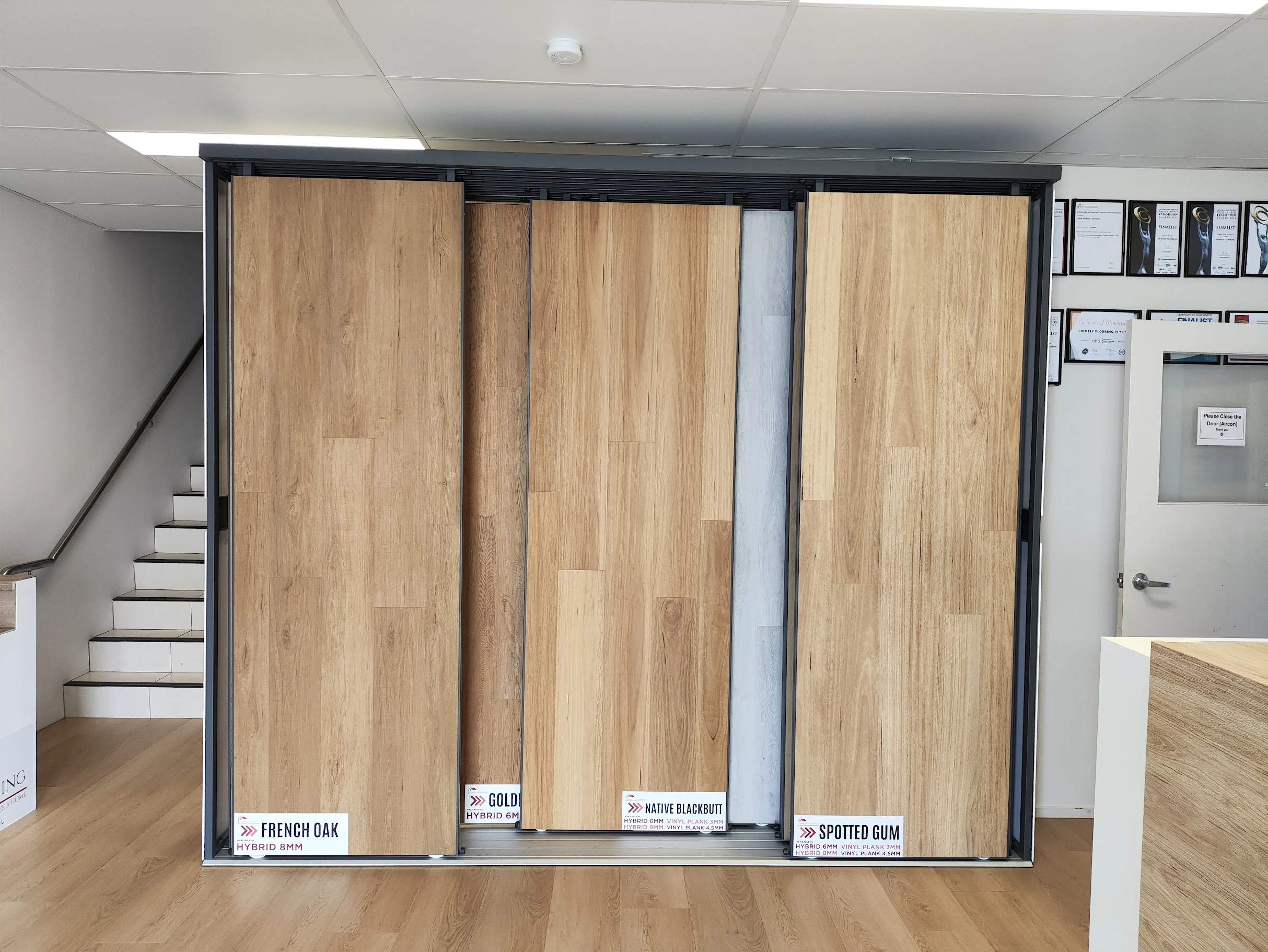 Interior of a flooring showroom featuring large sliding display racks of light wood flooring samples. A text overlay at the bottom reads: 'ON THE BLOG — HYBRID VS. LAMINATE VS. VINYL: WHICH IS THE BEST VALUE FOR QUEENSLAND HOMES?